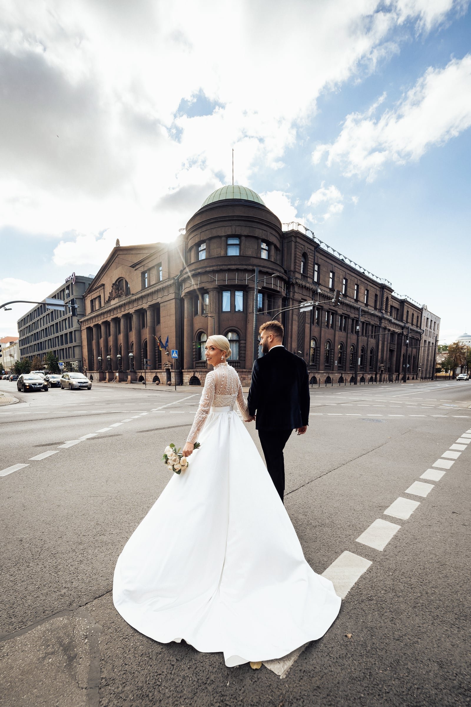 Bride looking back over her shoulder as she and the groom cross an empty street towards a domed interwar stone building in central Kaunas