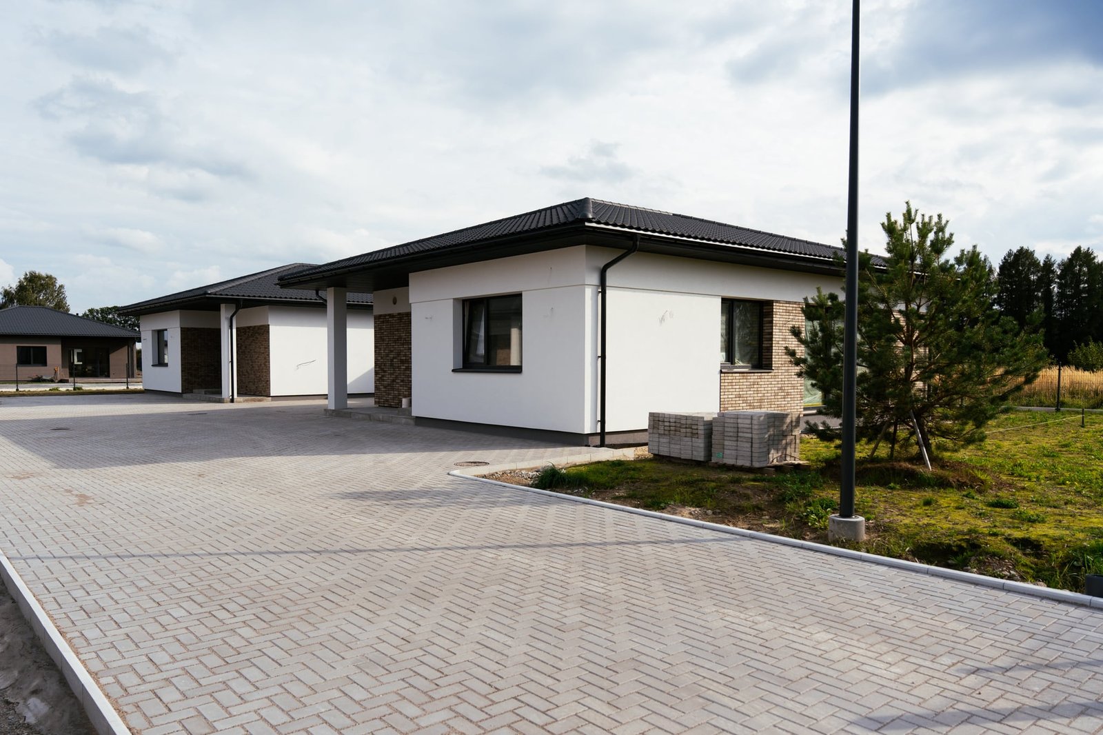 Kvartalo gatvė su eglučių raštu klotomis trinkelėmis, vėliavos stiebu ir baltais namais — prieš retušą