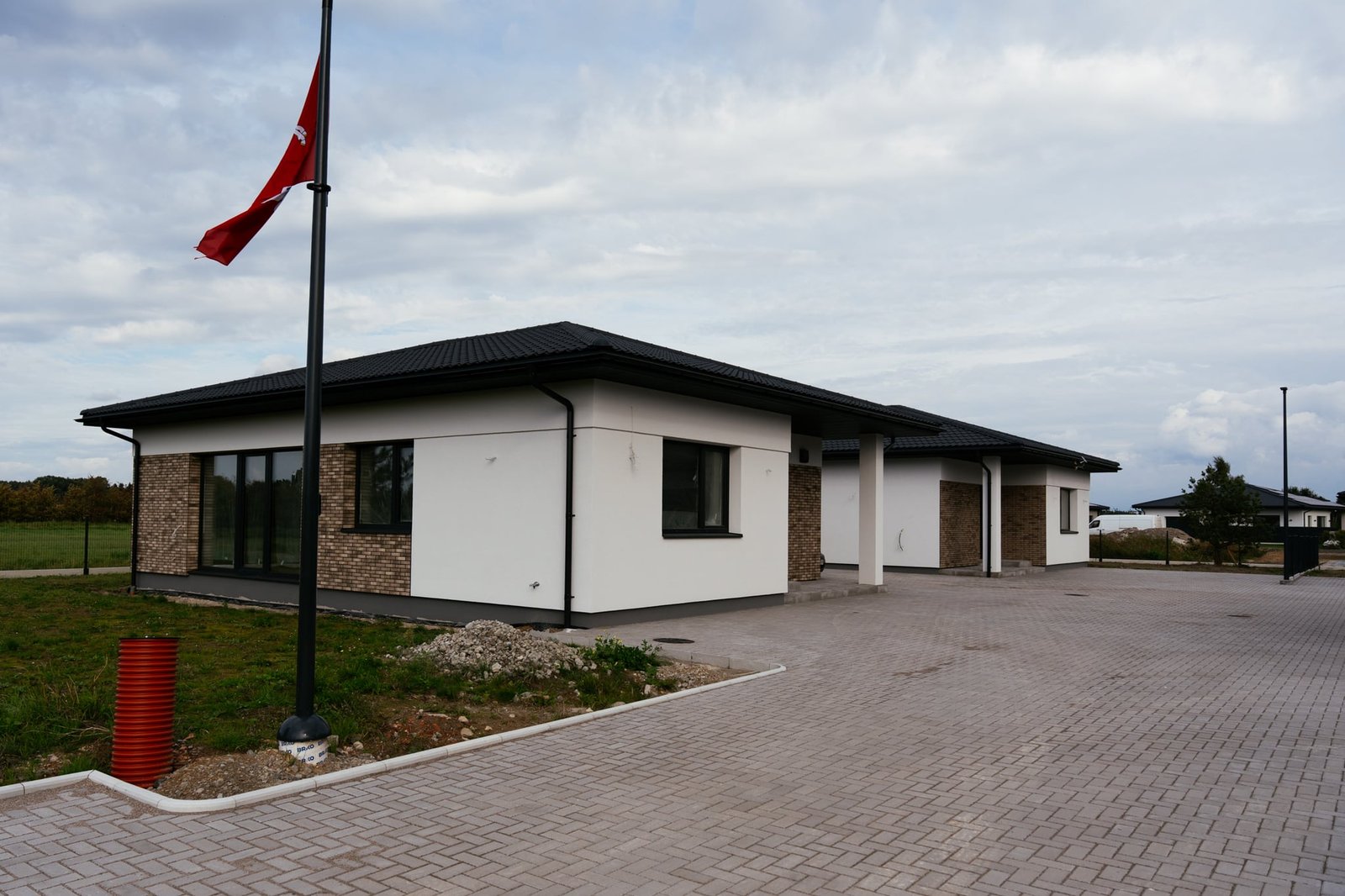 Baltas namas su plytų apdaila ir terasos stiklinėmis durimis, priešais vėliavos stiebas — prieš retušą